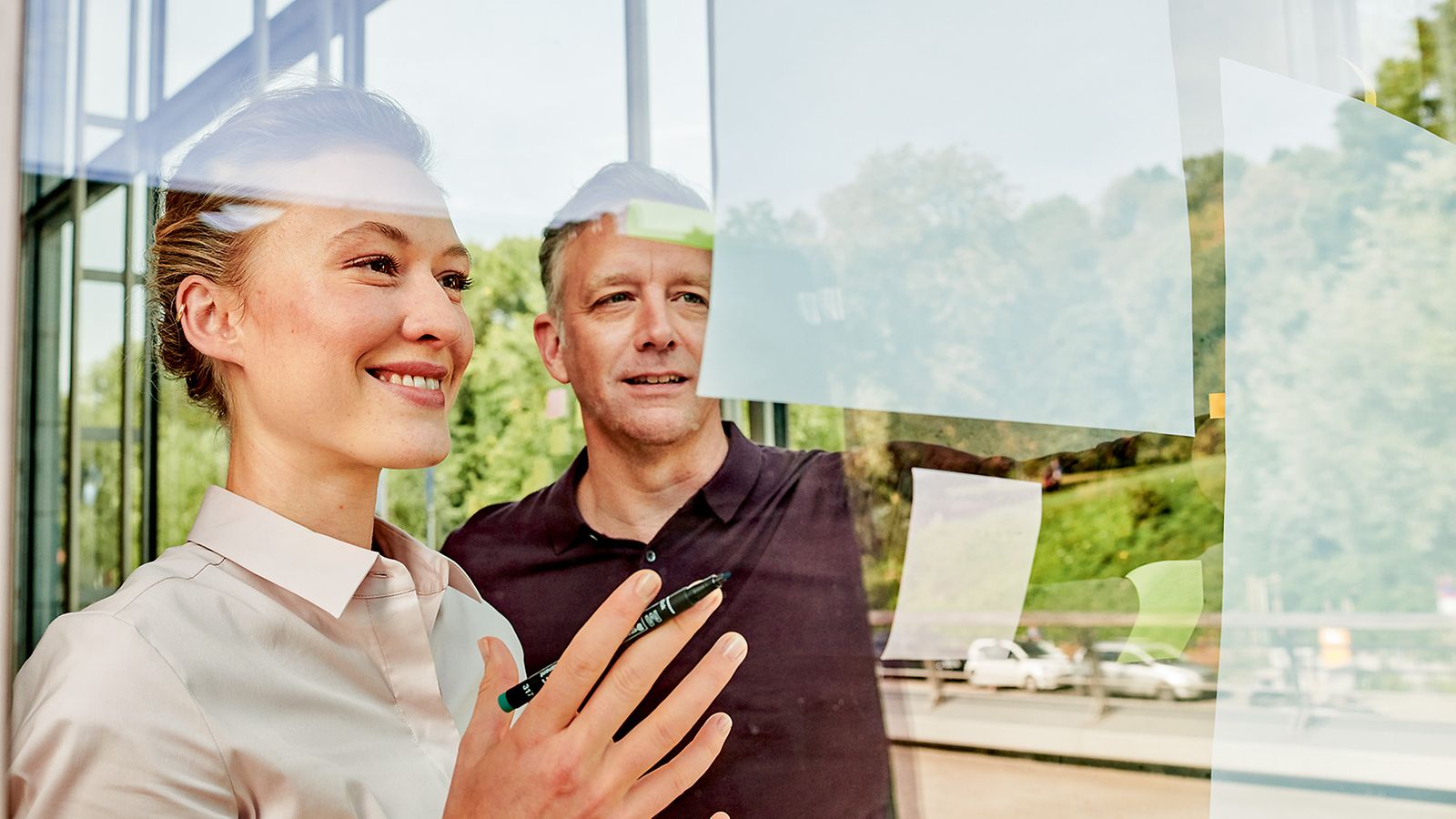 Eine Mann und eine Frau stehen vor einer Glaswand und zeigen freudig auf Pläne und Notizen an der Glaswand