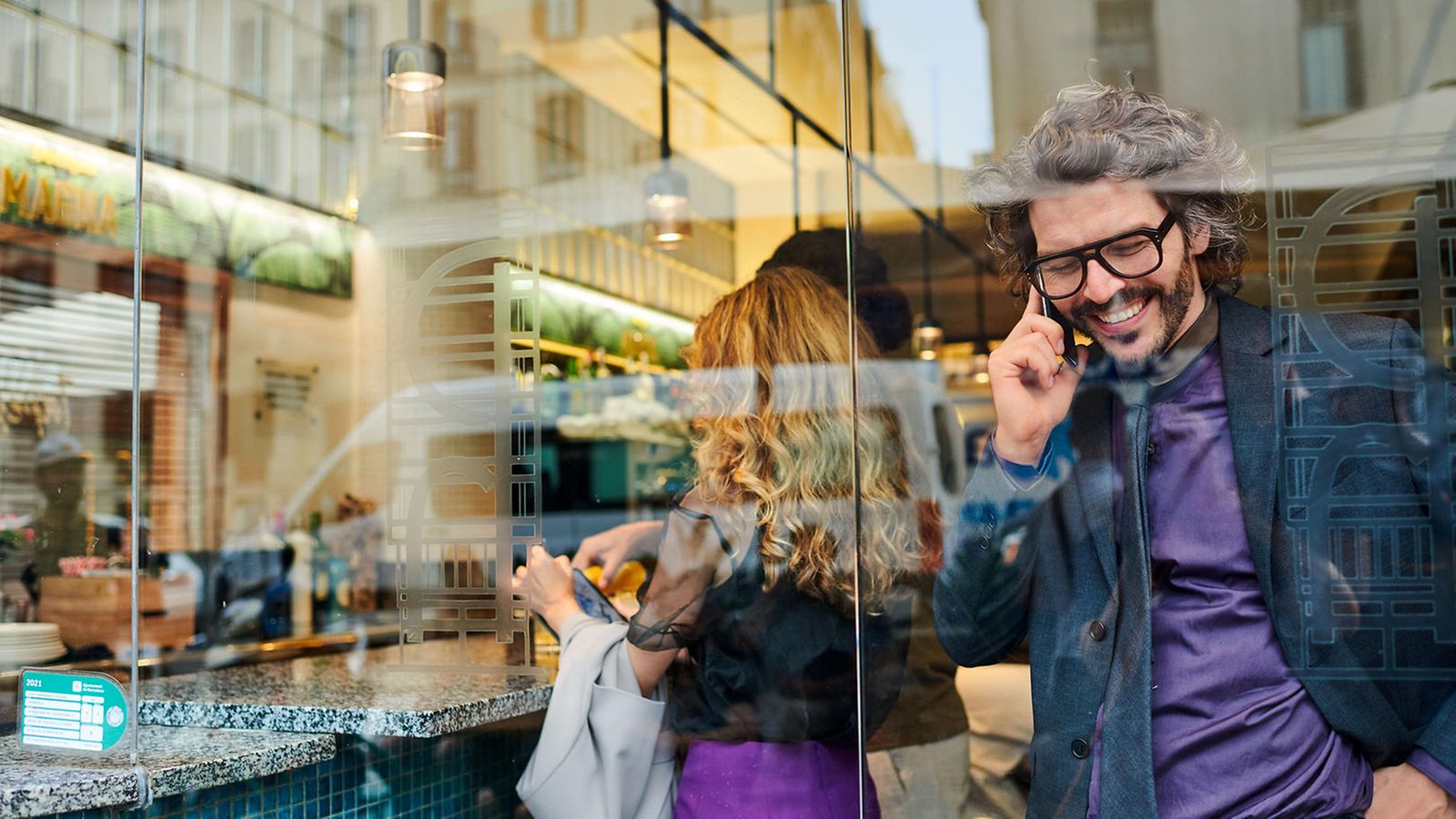 Behind a pane of glass, a man stands in the café and talks on the phone, smiling