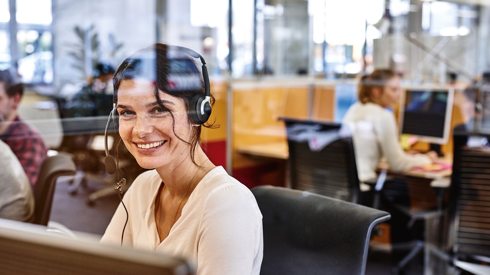 A service employee sits behind a pane of glass at her workplace with headphones and smiles into the camera