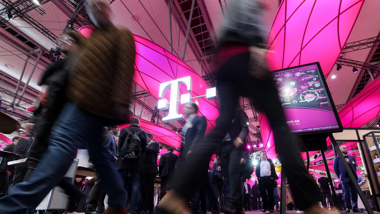 Blick nach oben vom Boden eines Events - rundherum viele Menschen (verschwommen), im Fokus das T-Logo an der magenta Decke