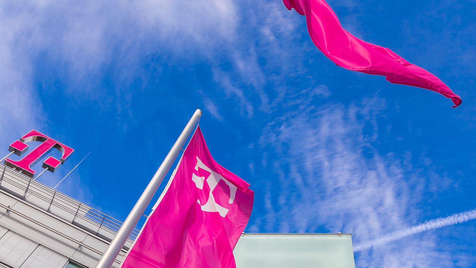 Blick in den blauen Himmel neben einem Gebäude mit magenta Telekom-Logo auf dem Dach und einer magenta Flage davor