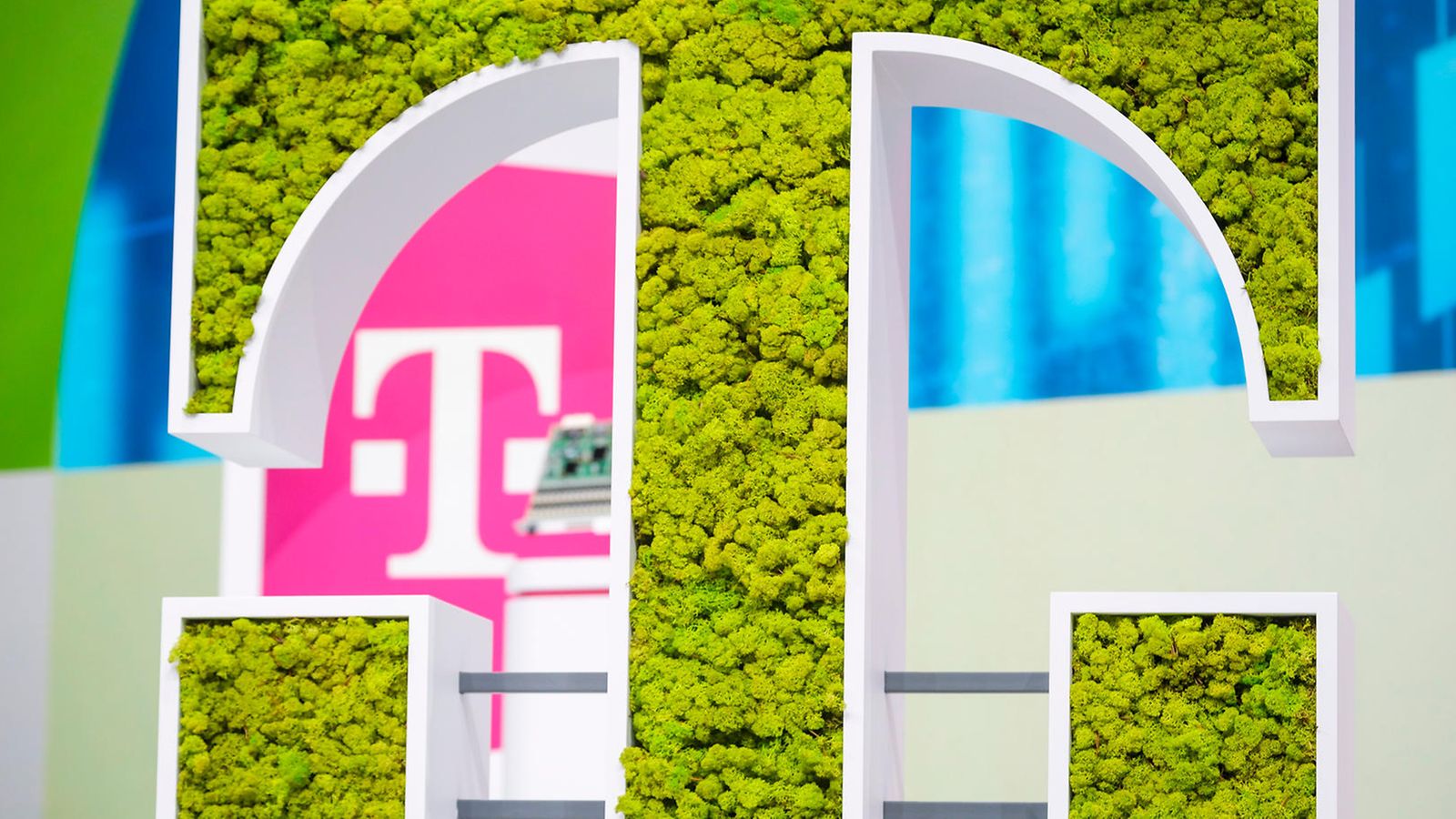 Ein großes grünes T-Logo mit Moos besetzt und im Hintergrund das T-Logo auf magenta Hintergrund